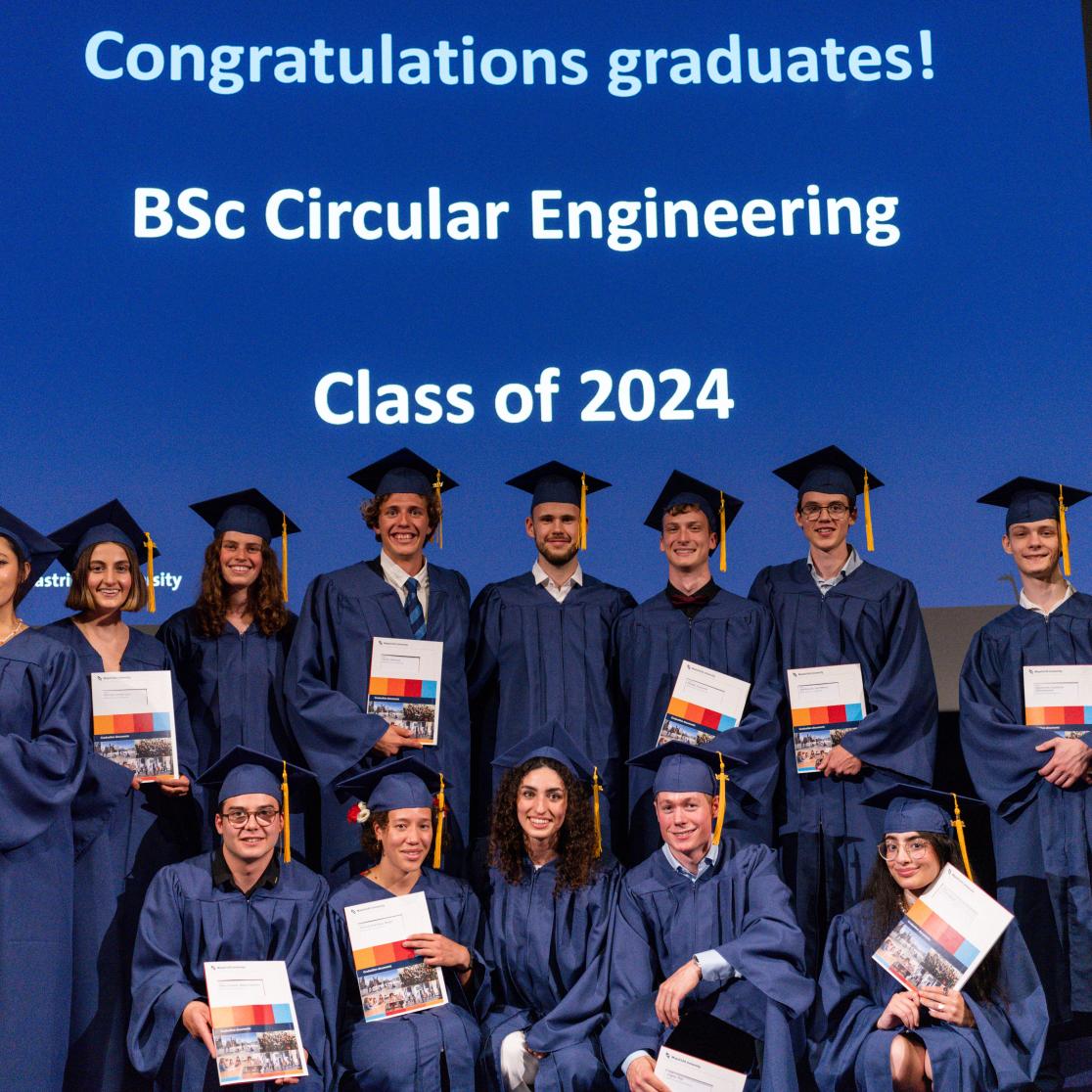 Thirteen students in blue gowns pose with their diploma in hands
