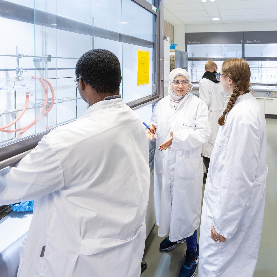 Multiple people in white coats and protective glasses discussing in a laboratory