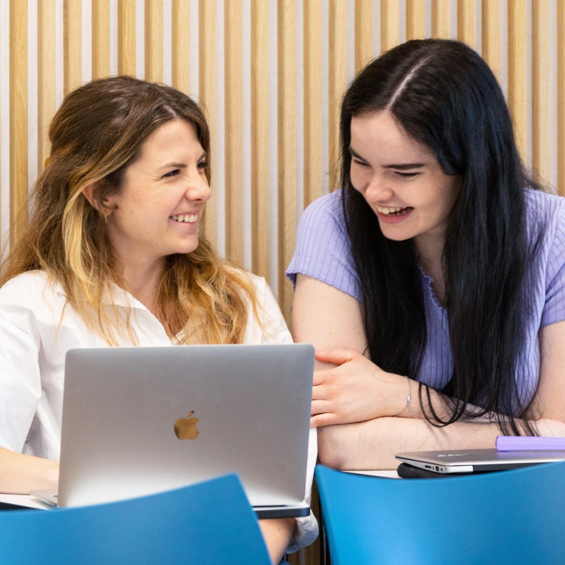 Two seated students with one laptop laughing.