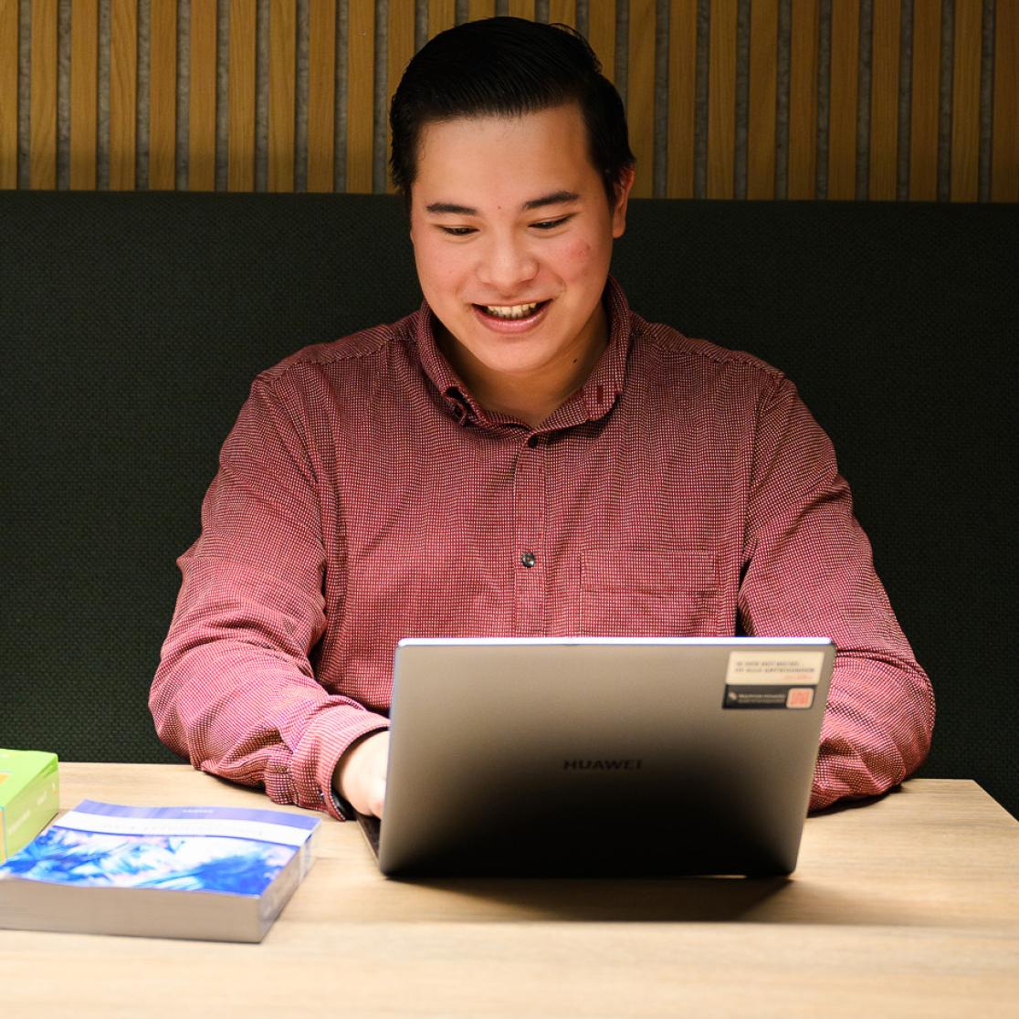 Student smiling while attending an online information session on a laptop, with books on the table in a university study area.