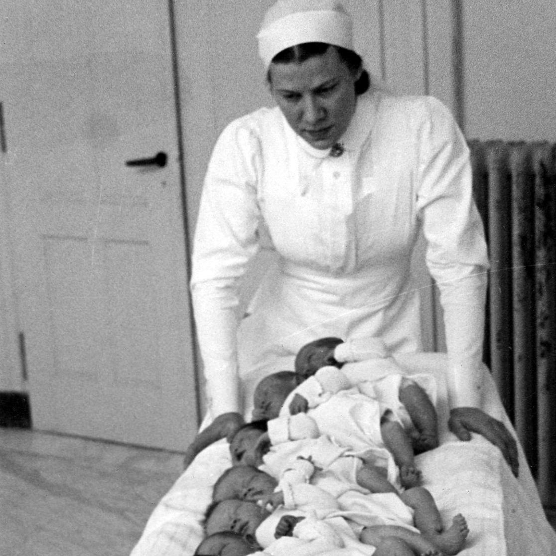 Tien zuigelingen op een bed. Zr A.M. Rulkens, hoofd verloskundige ziet toe. Heerlen, circa 1950.
