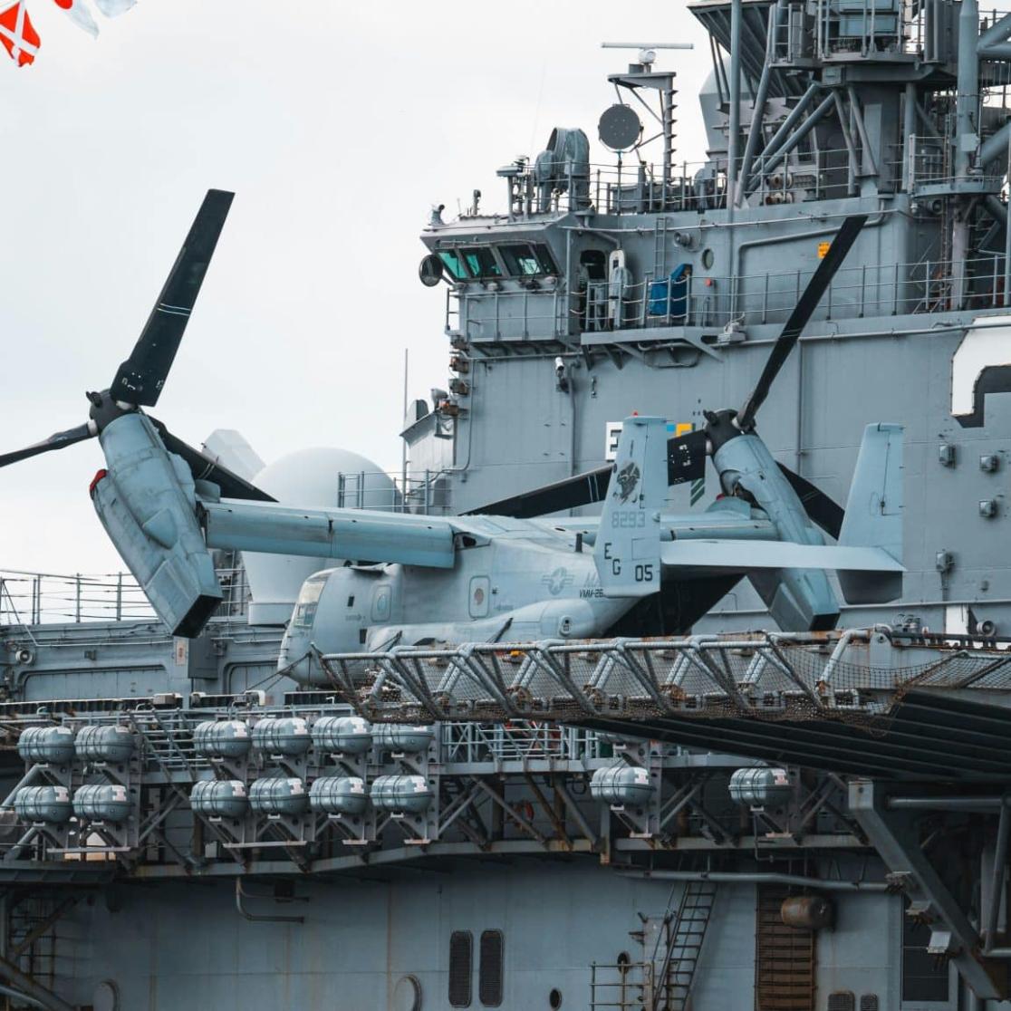 A U.S. Navy amphibious assault ship with a large number "3" on its side, carrying a V-22 Osprey tiltrotor aircraft and an AH-1Z Viper attack helicopter. The deck is lined with ordnance, and colorful signal flags are strung overhead.