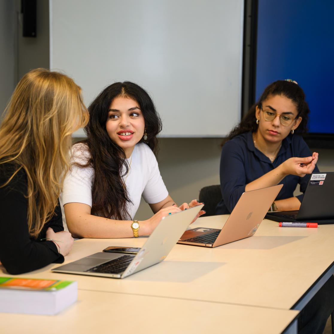 Drie studenten in gesprek tijdens een werkgroep, met laptops open op tafel.