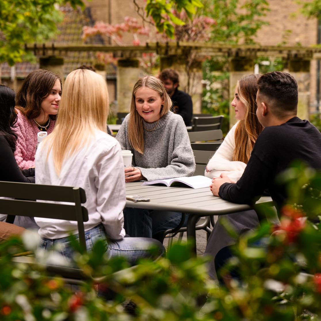 Een groep studenten zit samen aan een tafel in de faculteitstuin en bespreekt studiemateriaal in een informele sfeer.