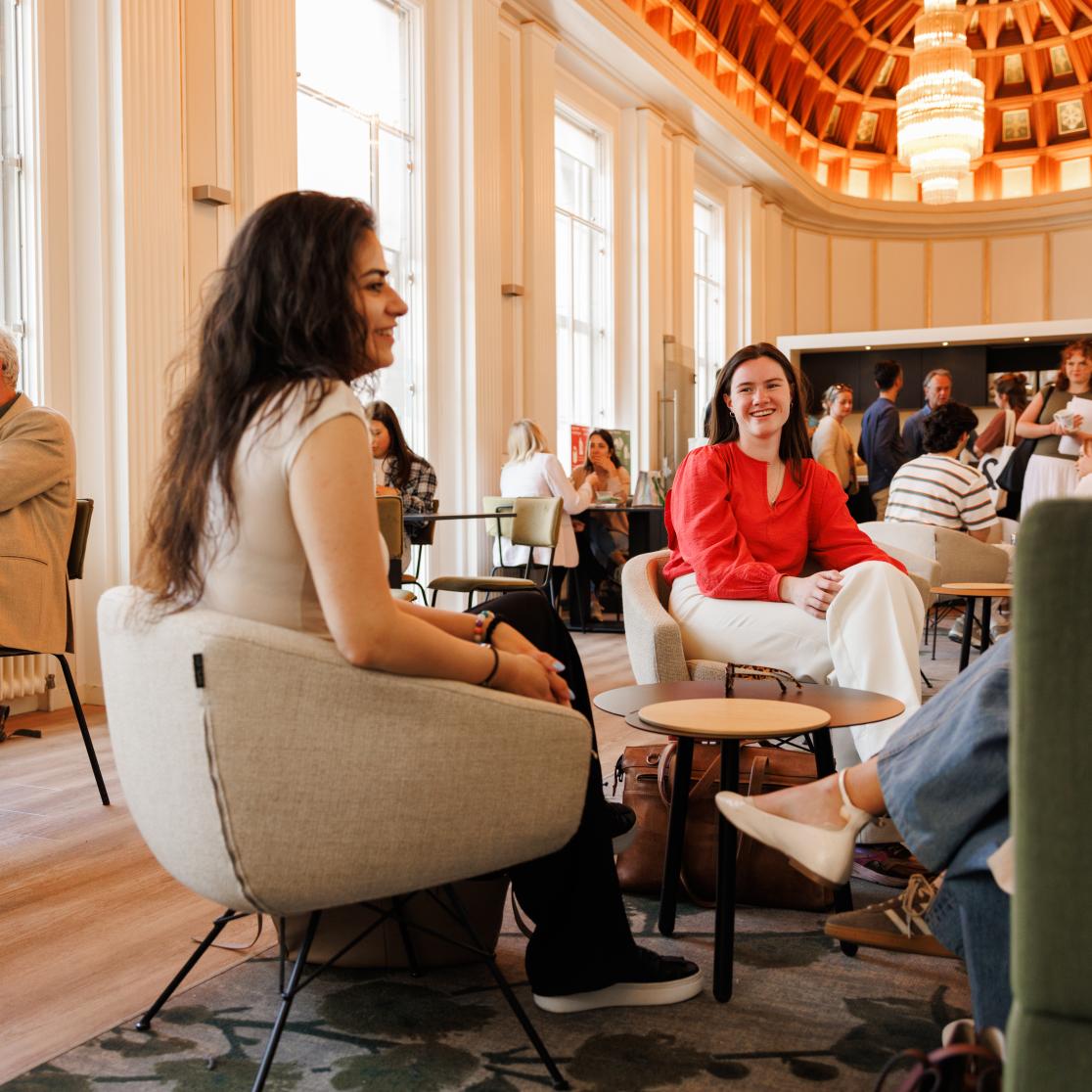 Studenten en staf in gesprek en ontspannen sfeer in de common room van de rechtenfaculteit, met hoge ramen, warme verlichting en een karakteristiek houten plafond.