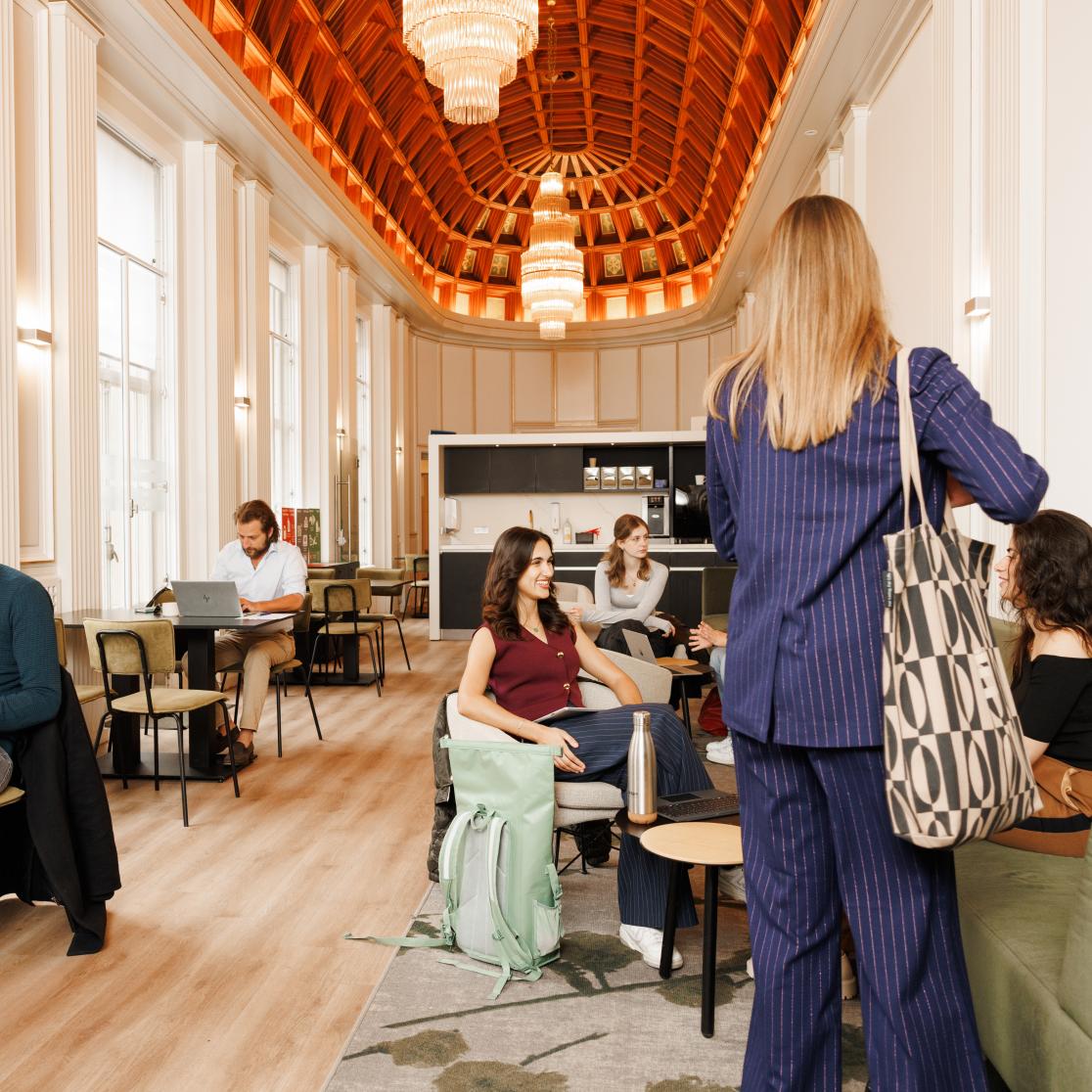 Studenten in gesprek, aan het lezen en werken in een lichte en sfeervolle common room met hoge ramen en houten plafond.