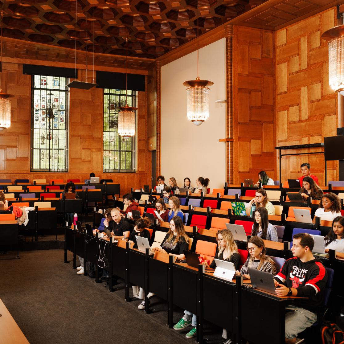 Lecture hall with wooden paneling, colorful seats, and stained glass windows. A professor speaks from a lectern while students listen attentively and work on laptops.