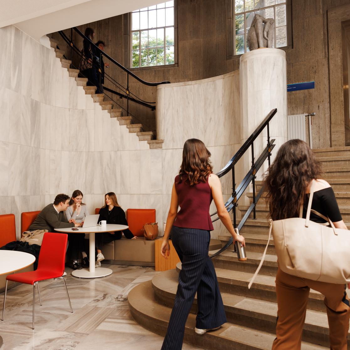 Students working and chatting in the marble hallway of the Lenculenhal, with seating along the wall and a grand staircase in view.