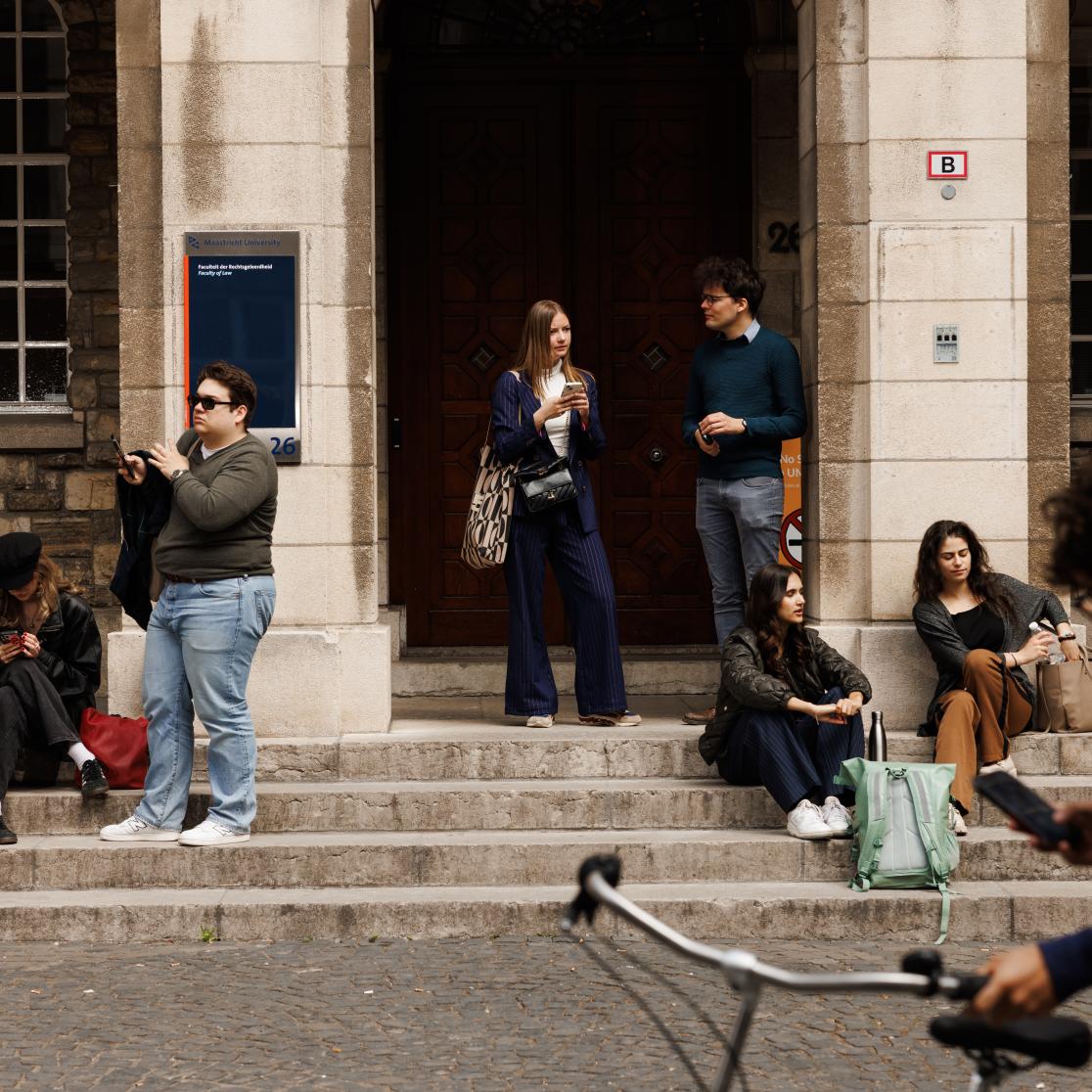 Students standing and sitting on the steps in front of the Faculty of Law entrance on Lenculenstraat, while a cyclist passes by in the foreground.