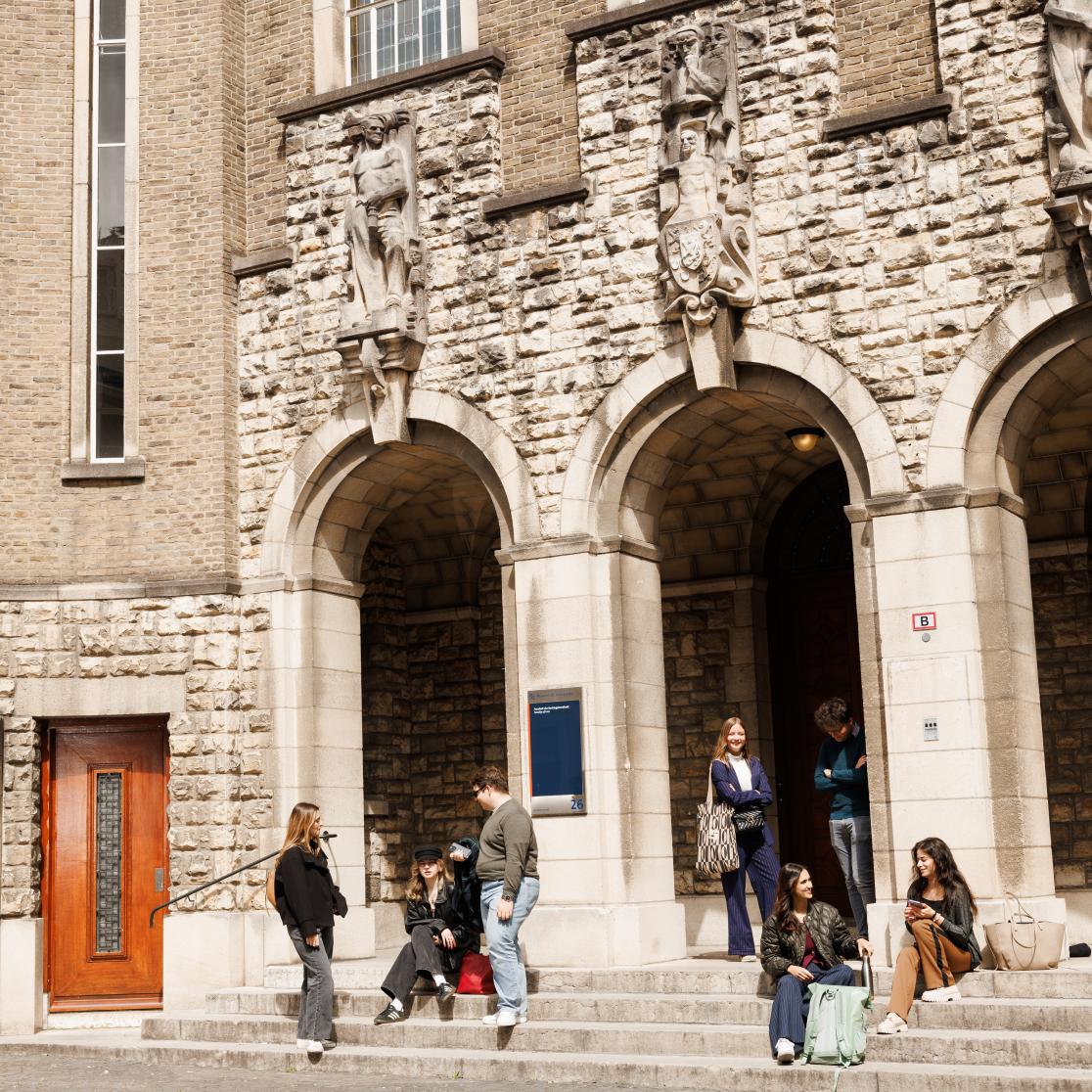 Students sitting and chatting on the steps in front of the monumental entrance of the Faculty of Law on Lenculenstraat in Maastricht, beneath sculpted figures on the facade.