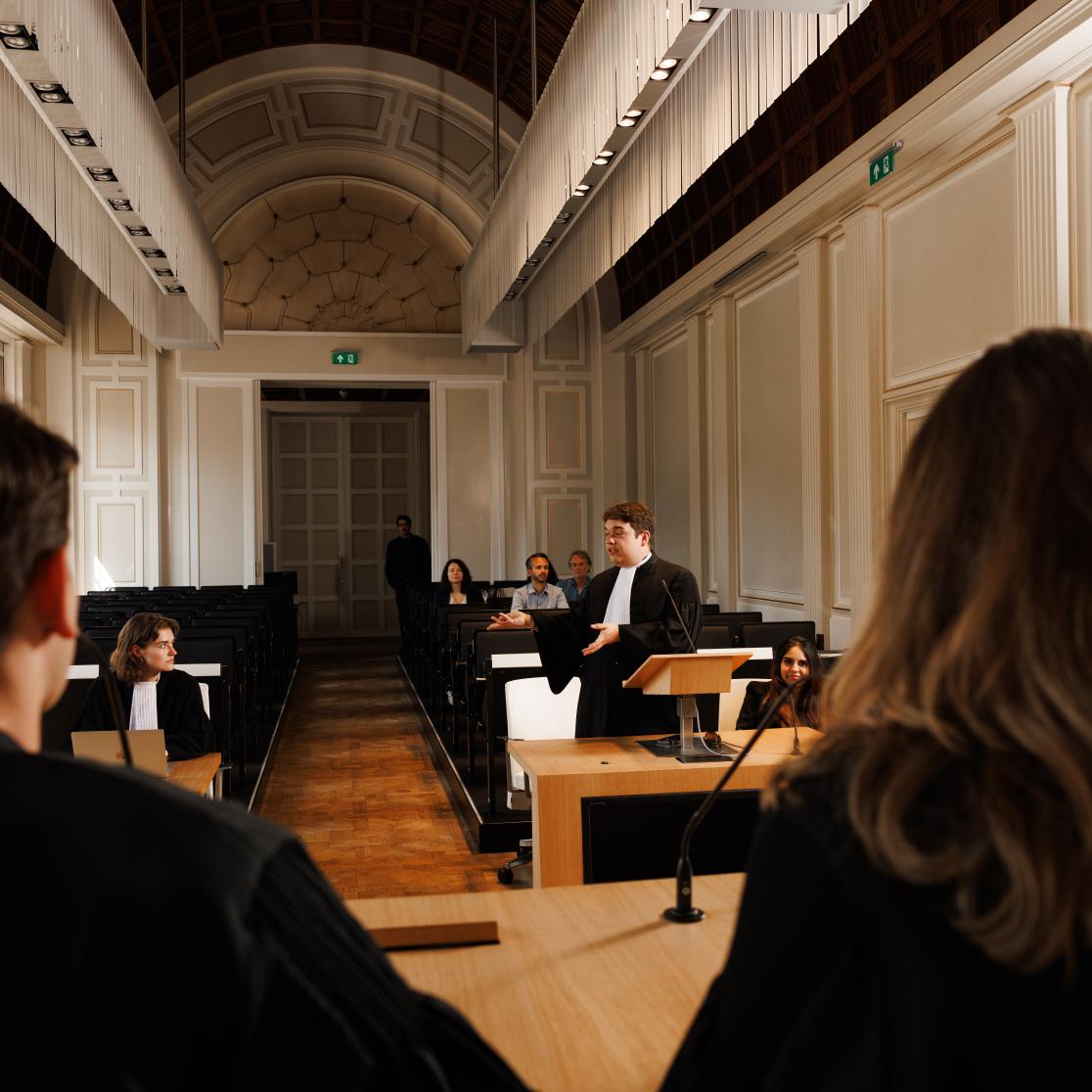 Student in toga houdt een pleidooi tijdens een moot court-sessie aan Maastricht University, met medestudenten en publiek in de rechtszaal.