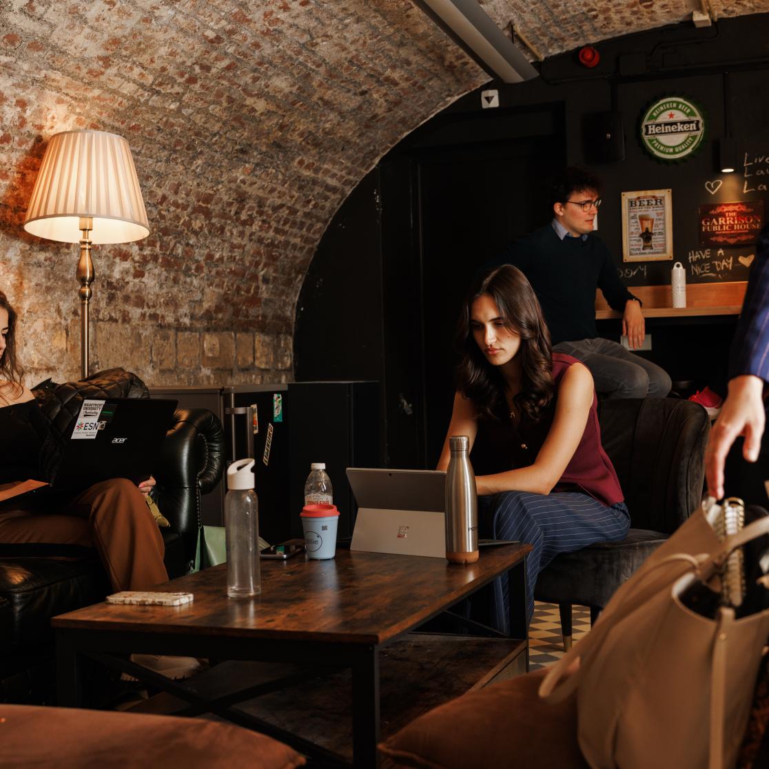 Students working on laptops in a cosy vaulted space with exposed brick walls, leather sofas, and a warm floor lamp; a bar area with posters and a chalkboard in the background.