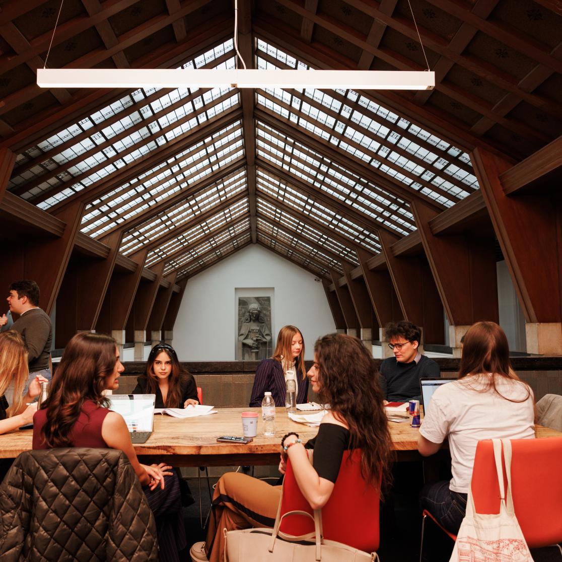 Students working together at a long wooden table beneath the iconic glass ceiling of the Faculty of Law, with the Lady Justice sculpture visible in the background.