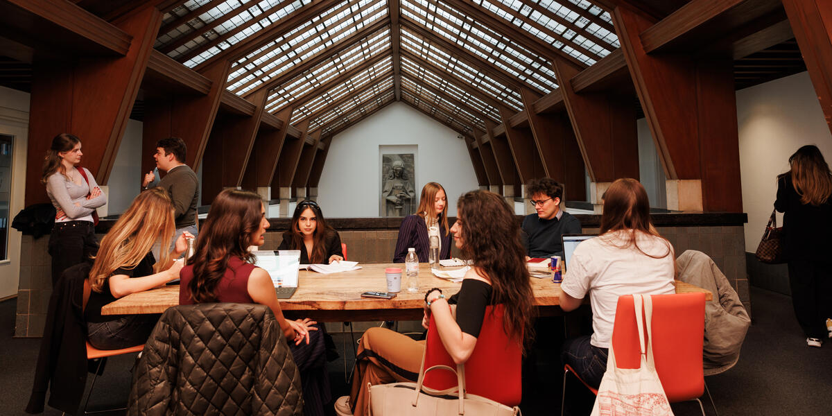 Students working together at a long wooden table beneath the iconic glass ceiling of the Faculty of Law, with the Lady Justice sculpture visible in the background.