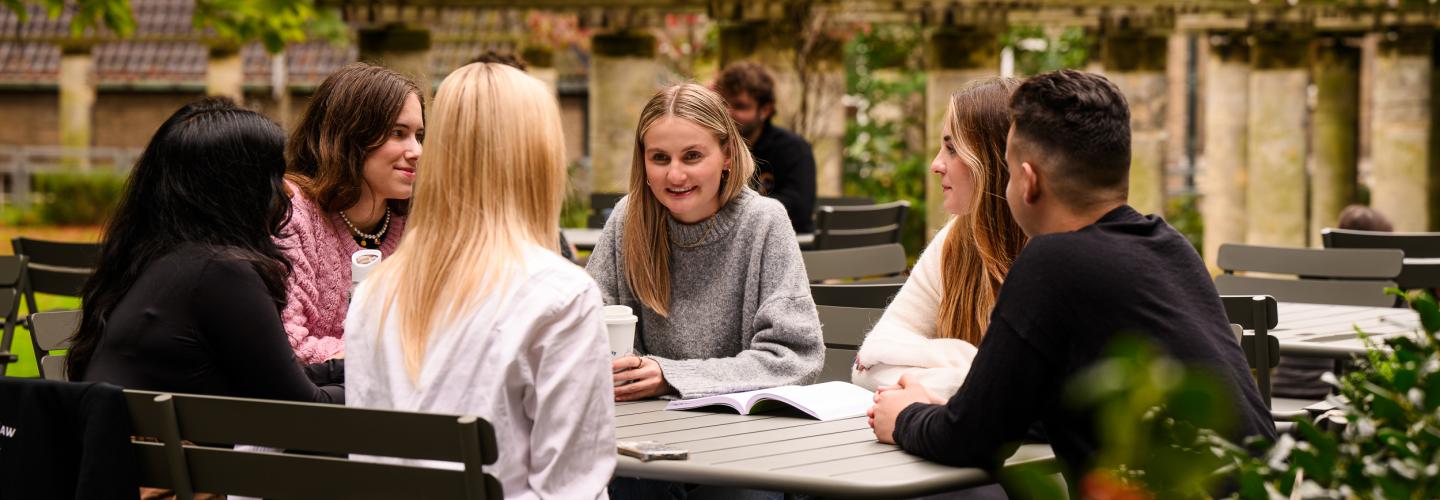 A group of students sits together at a table in the faculty garden, informally discussing study materials.