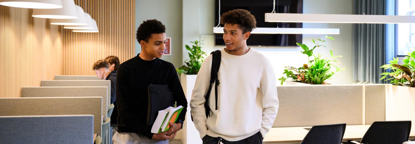 Two students walk through the faculty’s learning space while talking, with others studying in the background.