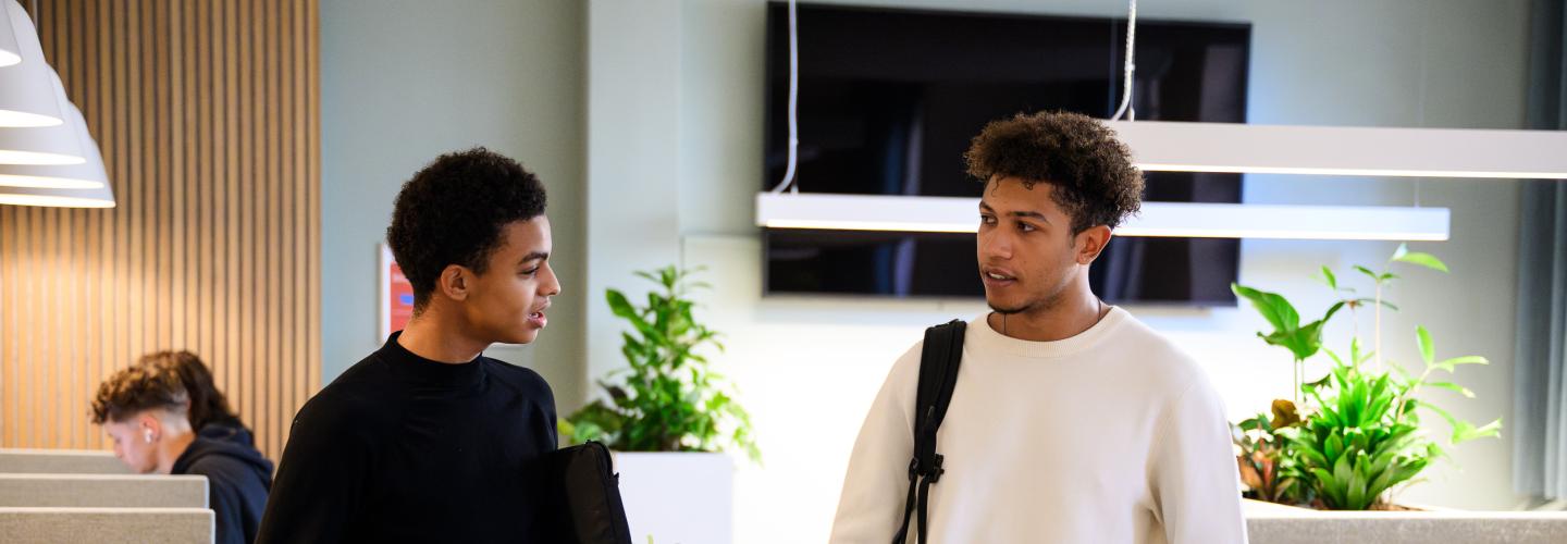 Twee studenten praten met elkaar in de learning space van de faculteit, met studieplekken op de achtergrond.
