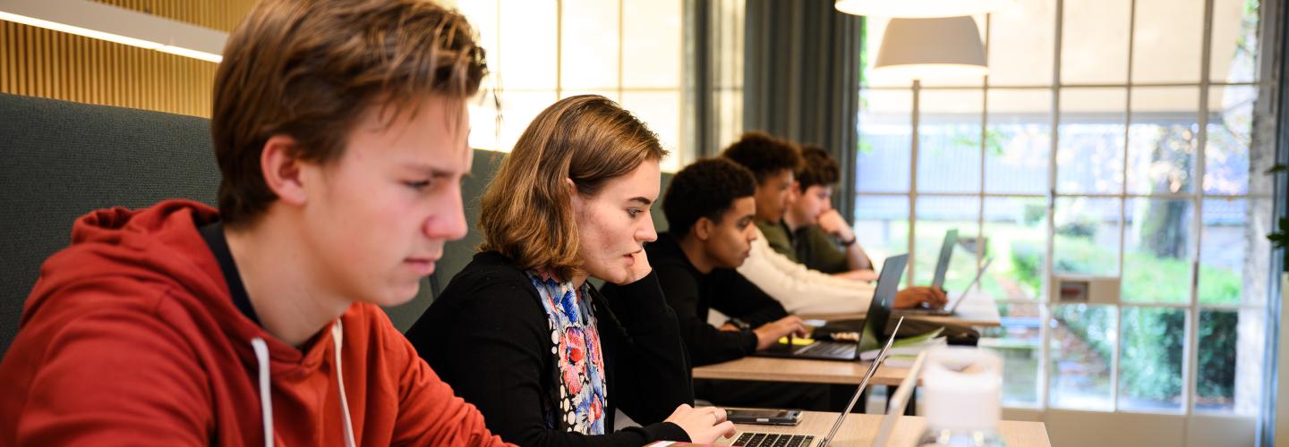 Studenten zitten naast elkaar aan lange tafels in de learning space en werken geconcentreerd aan hun laptop.