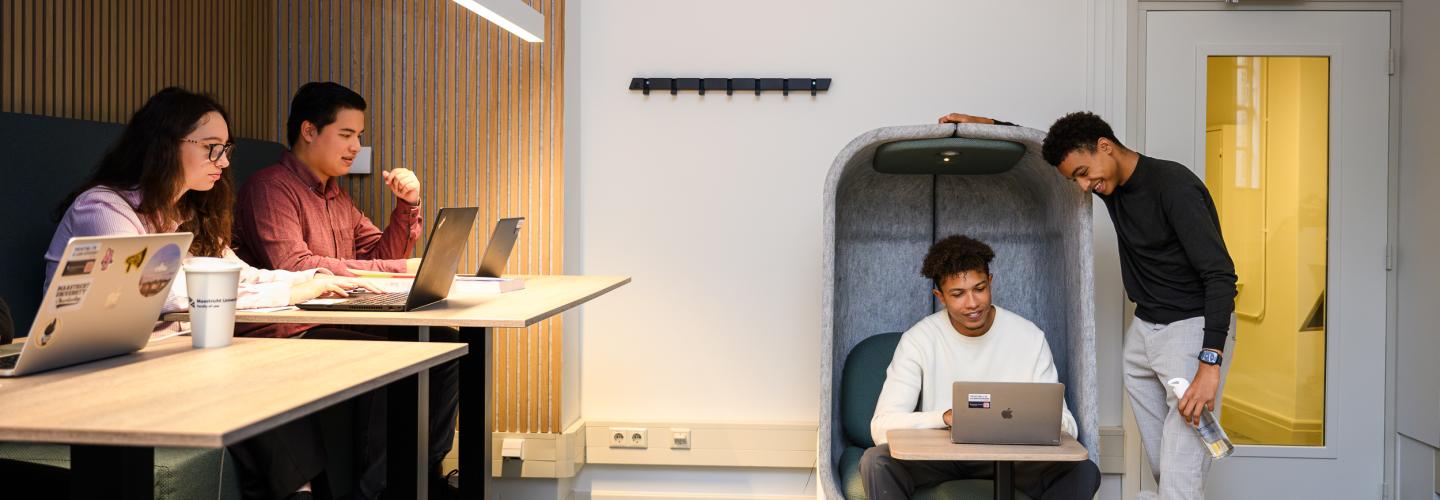 Twee studenten overleggen bij een laptop in een akoestische stoel, terwijl andere studenten geconcentreerd werken aan een tafel.