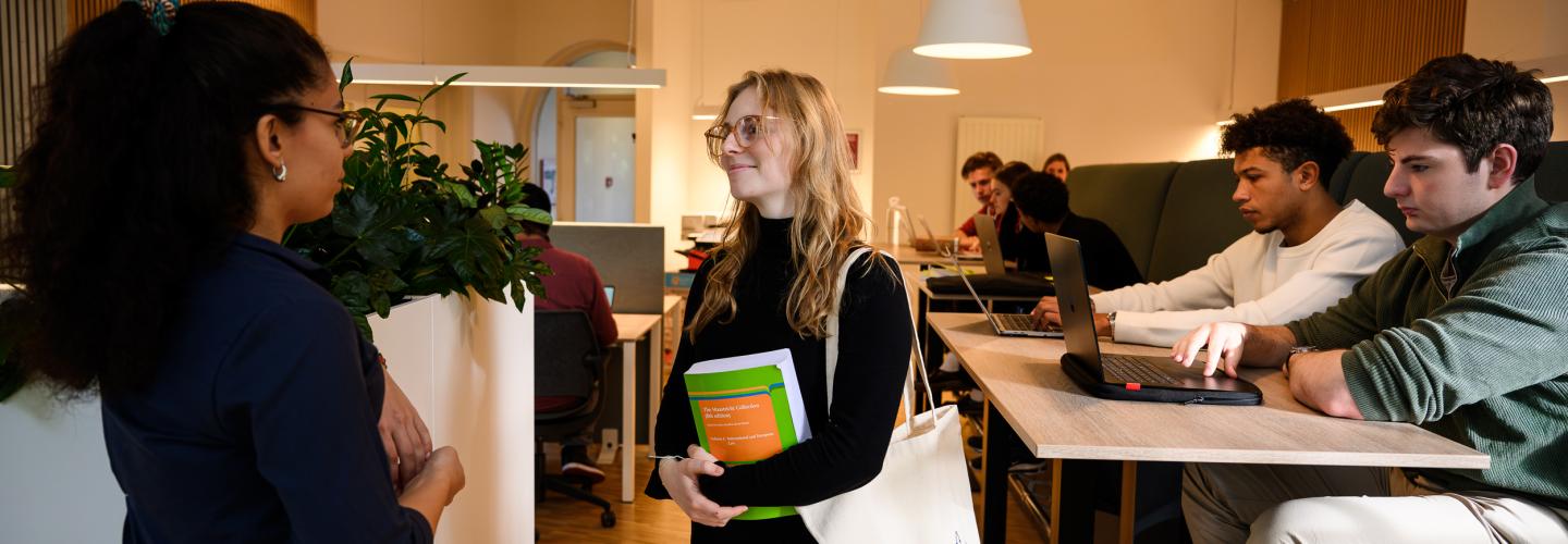 Twee studenten praten met elkaar in de learning space, terwijl anderen geconcentreerd werken achter hun laptops.
