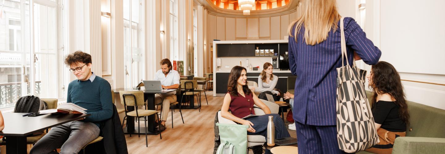 Studenten in gesprek, aan het lezen en werken in een lichte en sfeervolle common room met hoge ramen en houten plafond.