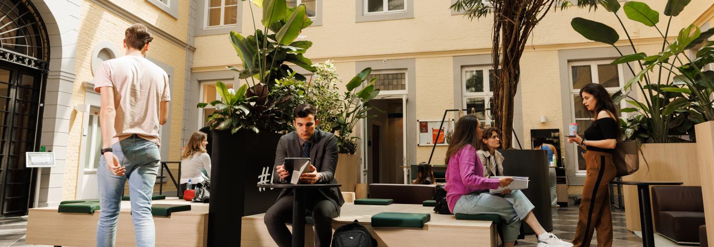 Students sitting and chatting in the green indoor courtyard of the Faculty of Law building on Kapoenstraat, surrounded by plants and natural light.