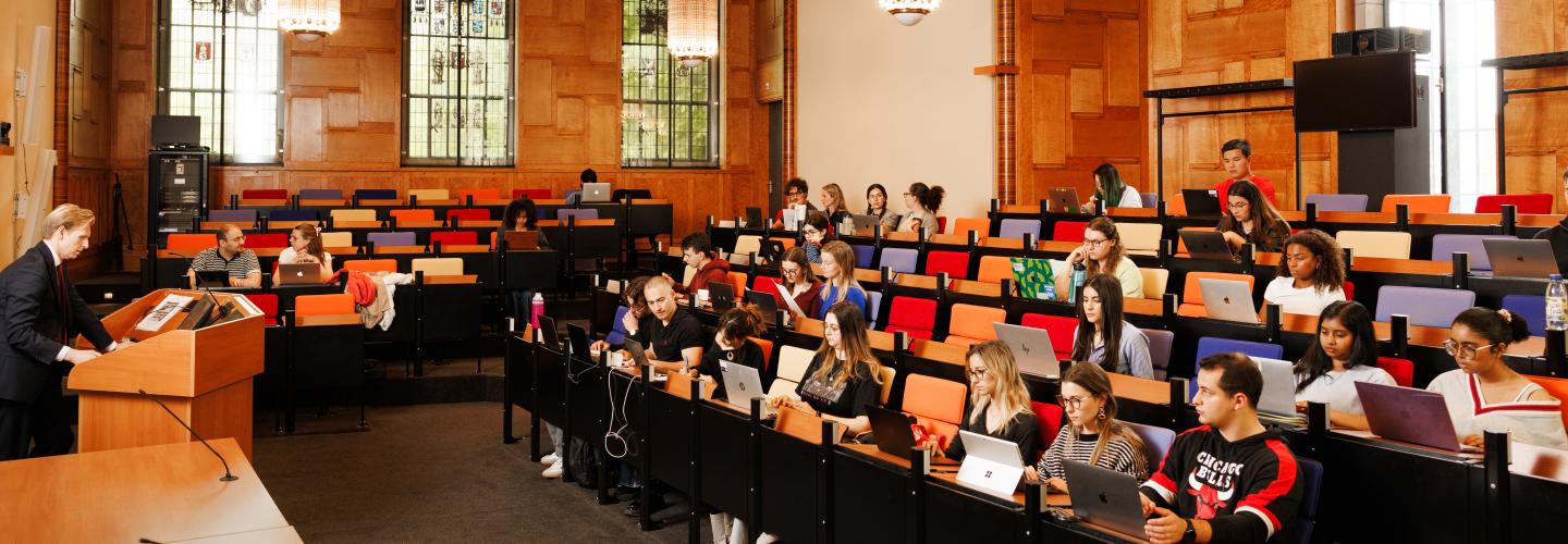 Lecture hall with wooden paneling, colorful seats, and stained glass windows. A professor speaks from a lectern while students listen attentively and work on laptops.
