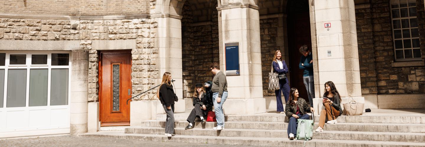 Students sitting and chatting on the steps in front of the monumental entrance of the Faculty of Law on Lenculenstraat in Maastricht, beneath sculpted figures on the facade.
