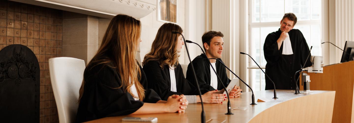Rechtenstudenten in toga voeren een oefenrechtszaak uit tijdens een moot court-sessie aan Maastricht University, terwijl een docent of begeleider toekijkt.