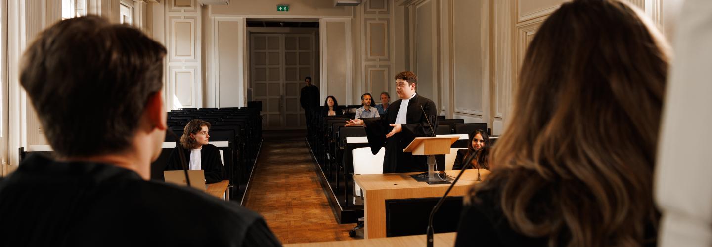 Student in toga houdt een pleidooi tijdens een moot court-sessie aan Maastricht University, met medestudenten en publiek in de rechtszaal.