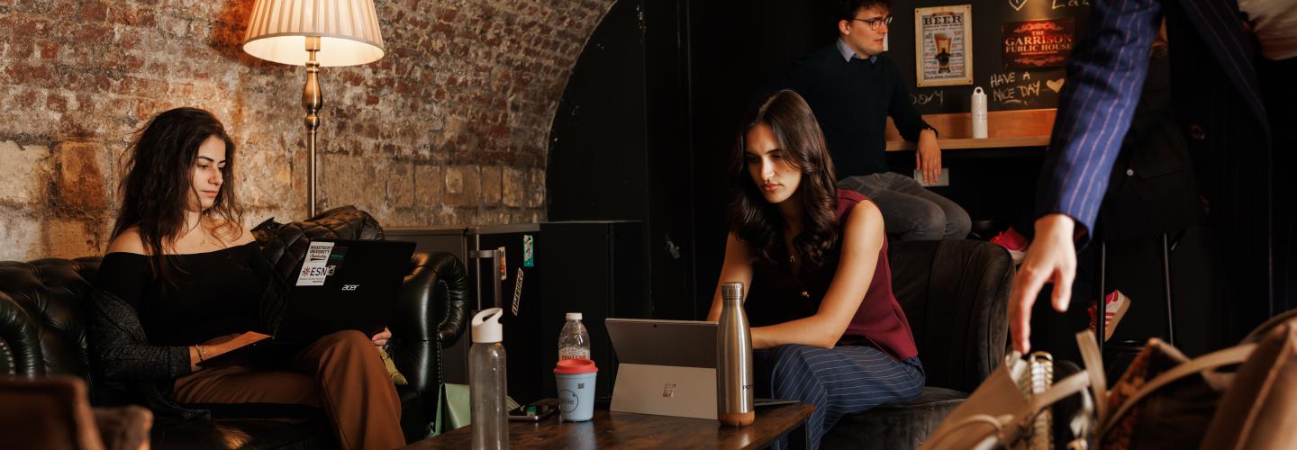 Students working on laptops in a cosy vaulted space with exposed brick walls, leather sofas, and a warm floor lamp; a bar area with posters and a chalkboard in the background.