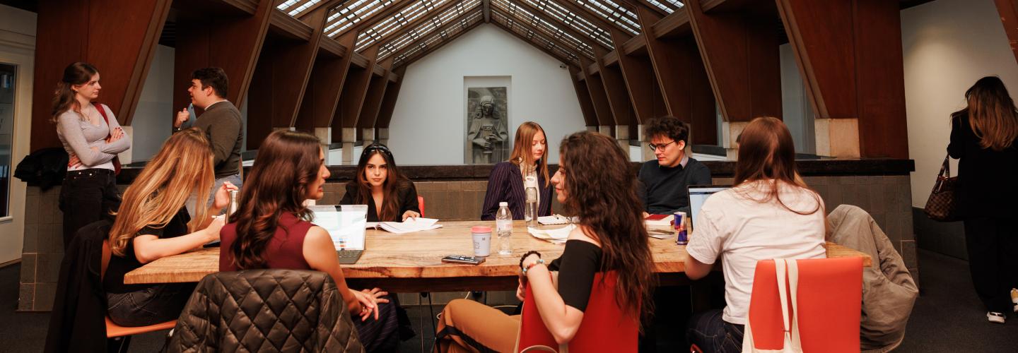 Students working together at a long wooden table beneath the iconic glass ceiling of the Faculty of Law, with the Lady Justice sculpture visible in the background.