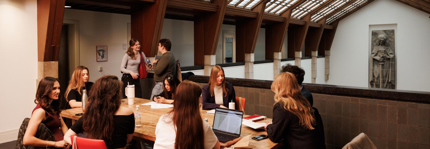 Studenten werken en overleggen aan een houten tafel onder een schuin glazen dak op de gang bij het standbeeld van Vrouwe Justitia in het gebouw van de rechtenfaculteit.