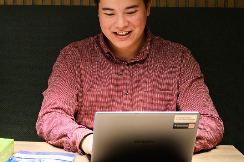 Student smiling while attending an online information session on a laptop, with books on the table in a university study area.