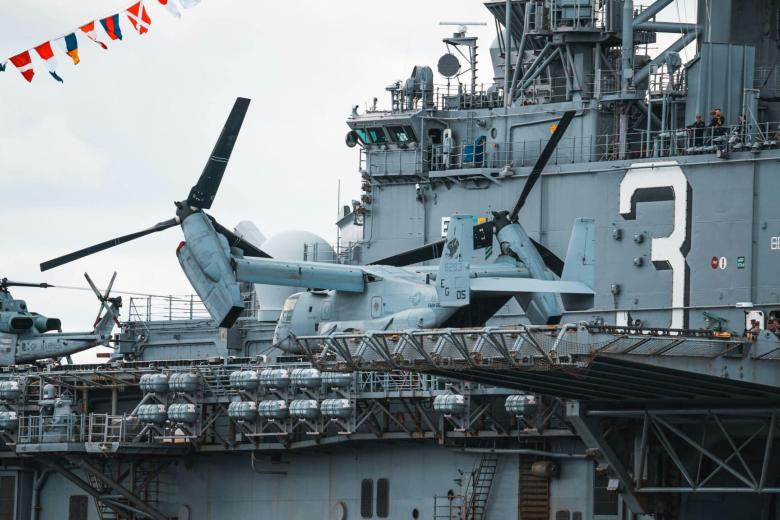A U.S. Navy amphibious assault ship with a large number "3" on its side, carrying a V-22 Osprey tiltrotor aircraft and an AH-1Z Viper attack helicopter. The deck is lined with ordnance, and colorful signal flags are strung overhead.