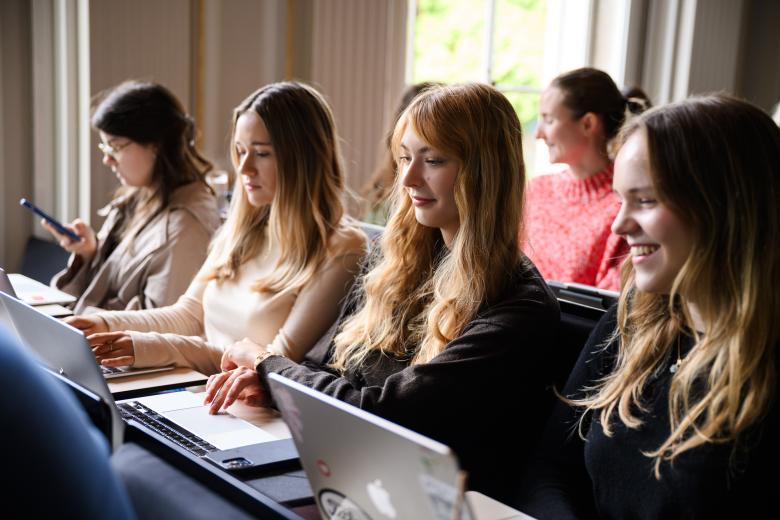 Students seated side by side in a lecture hall, working on their laptops during class.