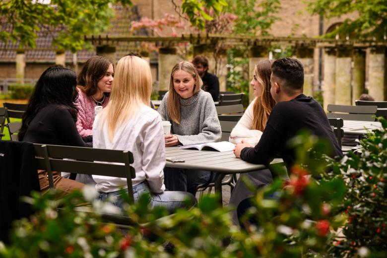 A group of students sits together at a table in the faculty garden, informally discussing study materials.