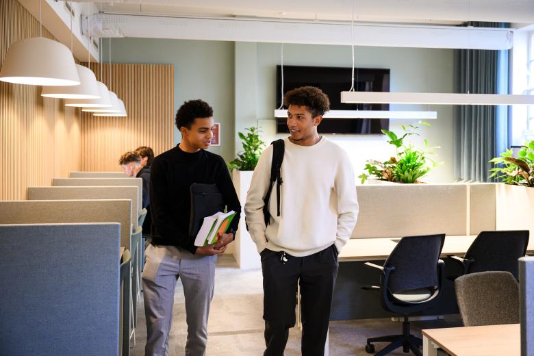 Twee studenten lopen door de learning space van de faculteit en praten met elkaar, terwijl anderen op de achtergrond studeren