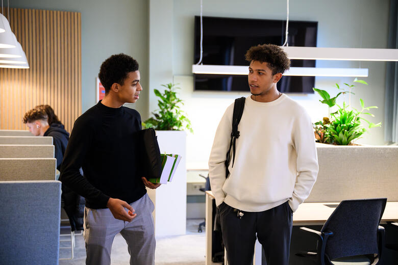 Twee studenten praten met elkaar in de learning space van de faculteit, met studieplekken op de achtergrond.