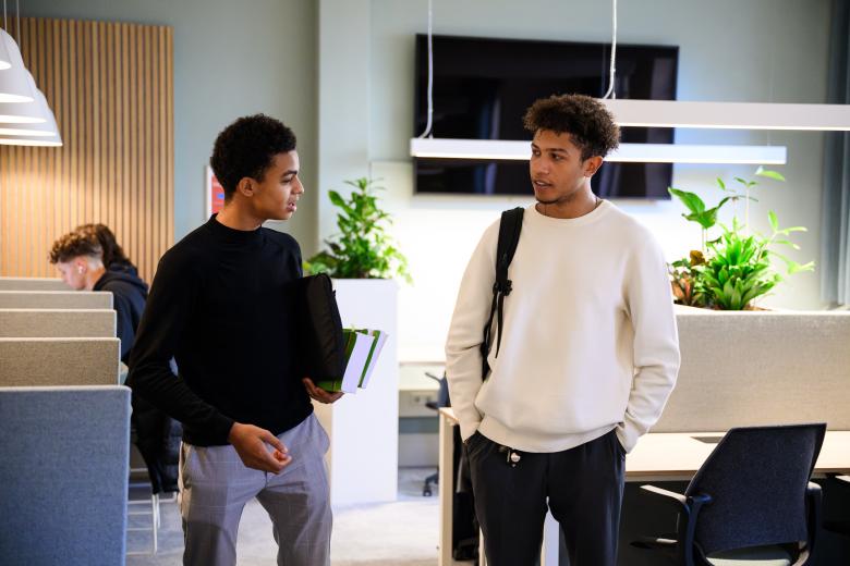 Twee studenten praten met elkaar in de learning space van de faculteit, met studieplekken op de achtergrond.