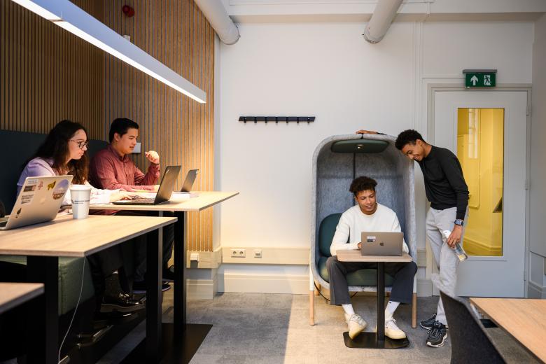 Twee studenten overleggen bij een laptop in een akoestische stoel, terwijl andere studenten geconcentreerd werken aan een tafel.