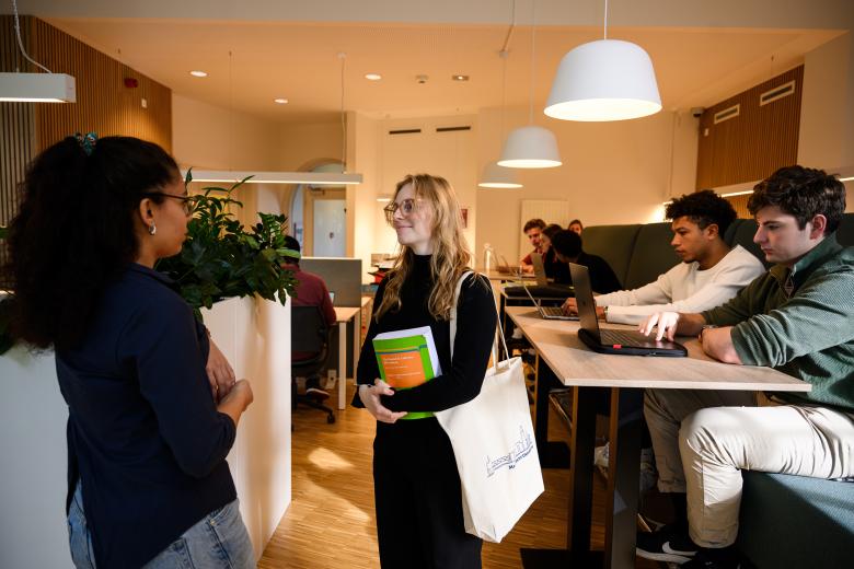 Twee studenten praten met elkaar in de learning space, terwijl anderen geconcentreerd werken achter hun laptops.