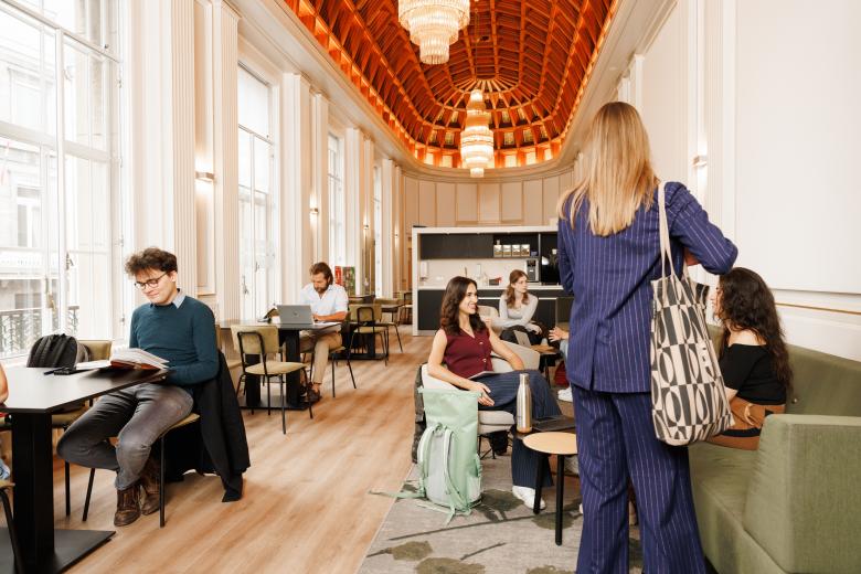 Studenten in gesprek, aan het lezen en werken in een lichte en sfeervolle common room met hoge ramen en houten plafond.