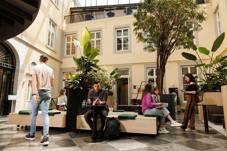 Students sitting and chatting in the green indoor courtyard of the Faculty of Law building on Kapoenstraat, surrounded by plants and natural light.