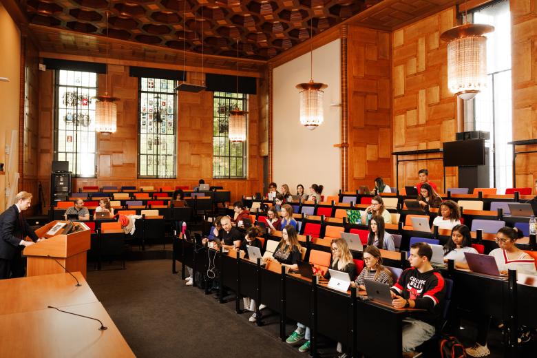 Lecture hall with wooden paneling, colorful seats, and stained glass windows. A professor speaks from a lectern while students listen attentively and work on laptops.