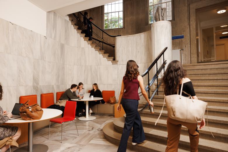 Students working and chatting in the marble hallway of the Lenculenhal, with seating along the wall and a grand staircase in view.