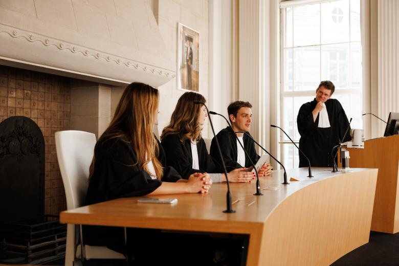 Rechtenstudenten in toga voeren een oefenrechtszaak uit tijdens een moot court-sessie aan Maastricht University, terwijl een docent of begeleider toekijkt.