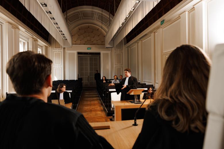 Student in toga houdt een pleidooi tijdens een moot court-sessie aan Maastricht University, met medestudenten en publiek in de rechtszaal.
