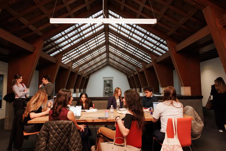 Students working together at a long wooden table beneath the iconic glass ceiling of the Faculty of Law, with the Lady Justice sculpture visible in the background.