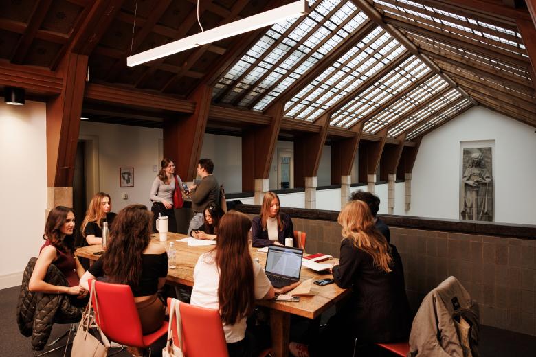 Studenten werken en overleggen aan een houten tafel onder een schuin glazen dak op de gang bij het standbeeld van Vrouwe Justitia in het gebouw van de rechtenfaculteit.